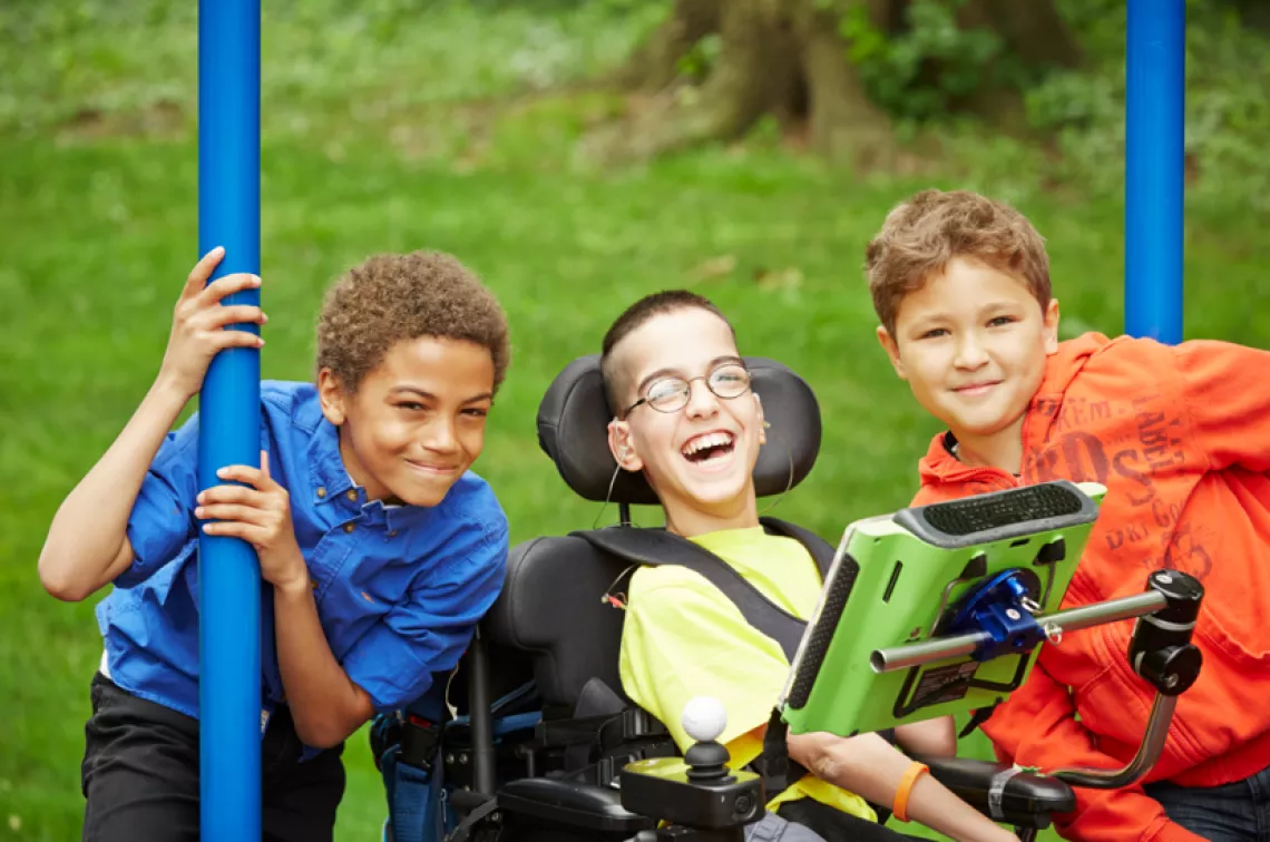 Three boys with one in a wheelchair smiling and using assistive device